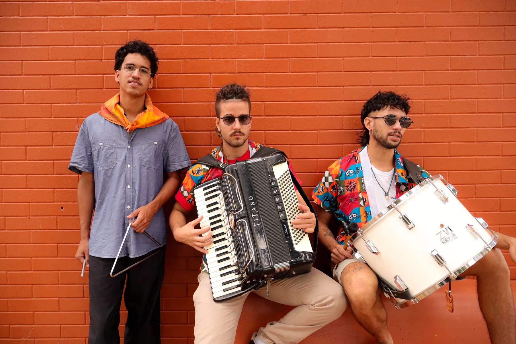O Trio Massapé diante de um muro de tijolos laranja, com triângulo, sanfona e zabumba